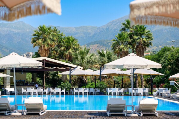 A pool area with lounge chairs and umbrellas, surrounded by palm trees and mountains in the background. - Rapos Resort Hotel (Himarë)