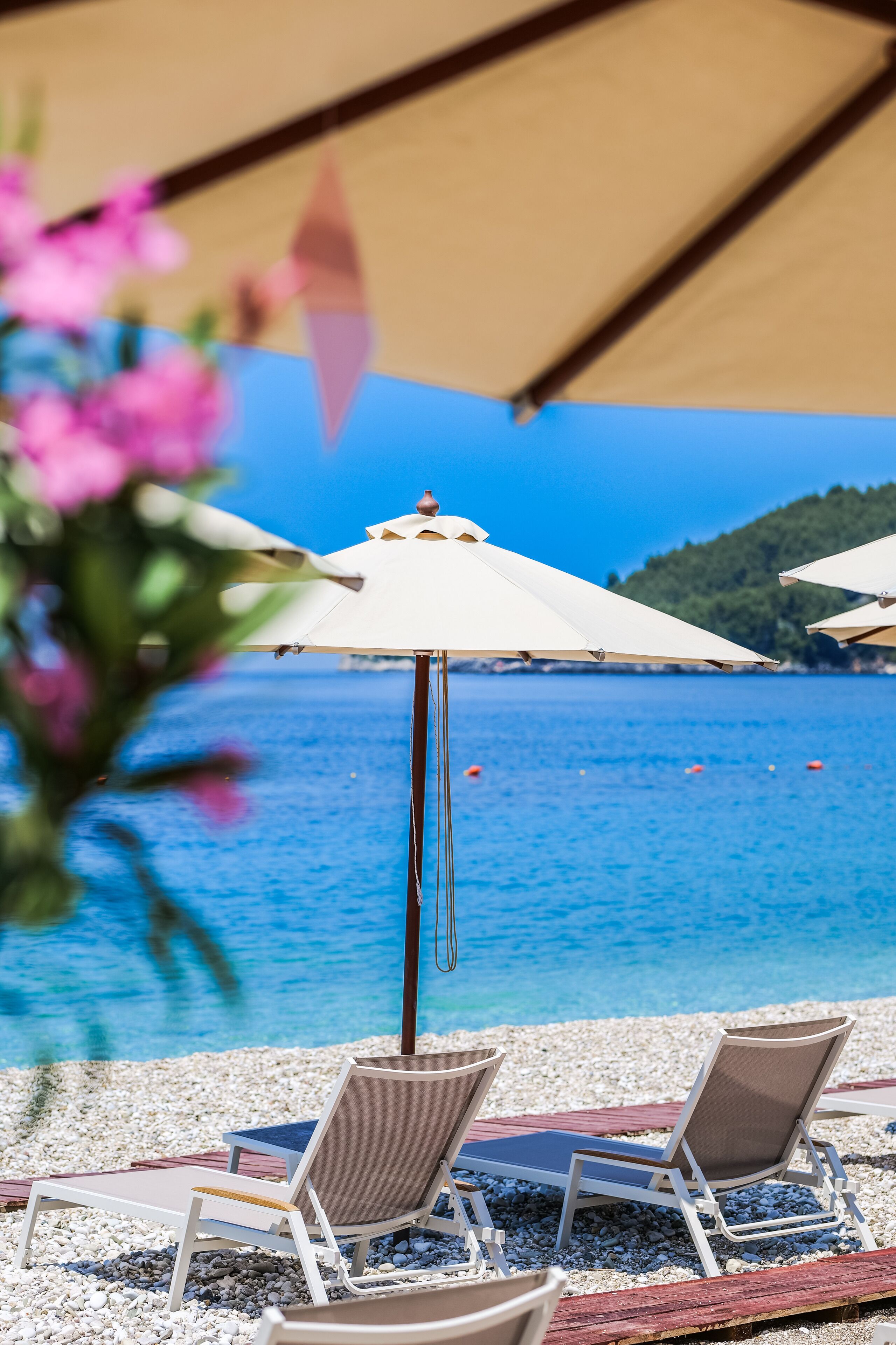 A beach with two sun loungers and a white umbrella, a pebble beach, and a clear blue sea.