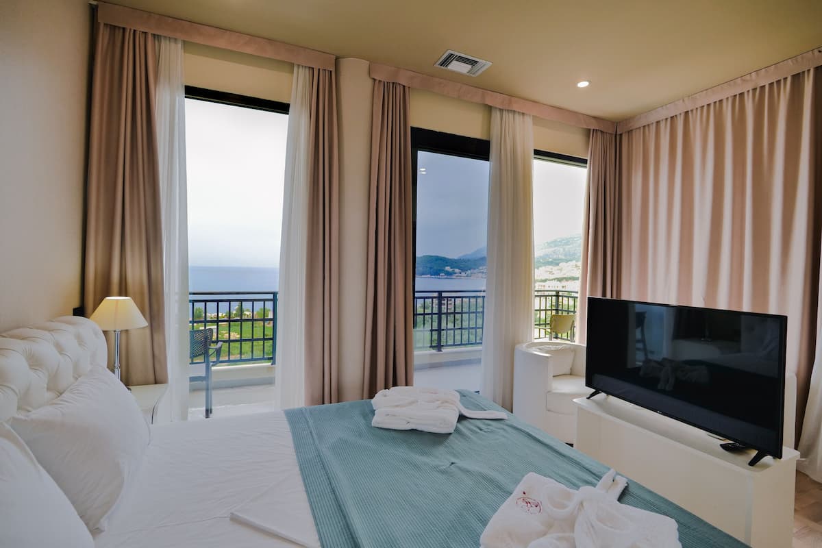 A hotel room with a large bed, a TV, and a balcony with a view of the sea.