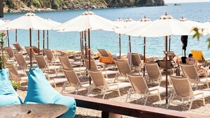 Un espacio de descanso en la playa con hileras de tumbonas y sombrillas, un banco de madera y una costa rocosa con una colina boscosa al fondo.