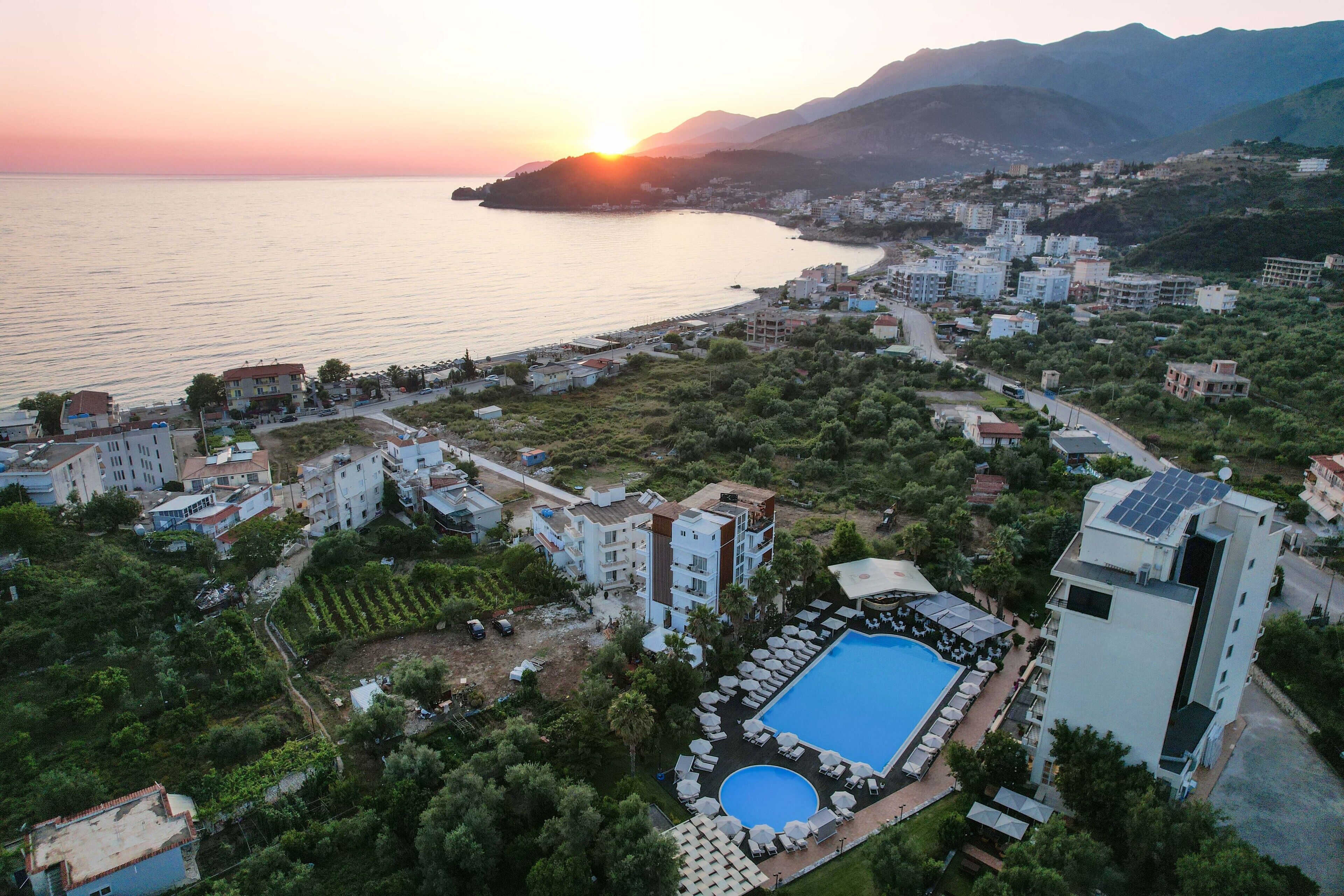 Vista aérea de un complejo turístico costero con piscina, varias construcciones y un atardecer sobre el mar.
