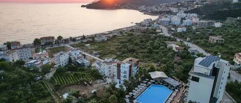 Vista aérea de un complejo turístico costero con piscina, varias construcciones y un atardecer sobre el mar.