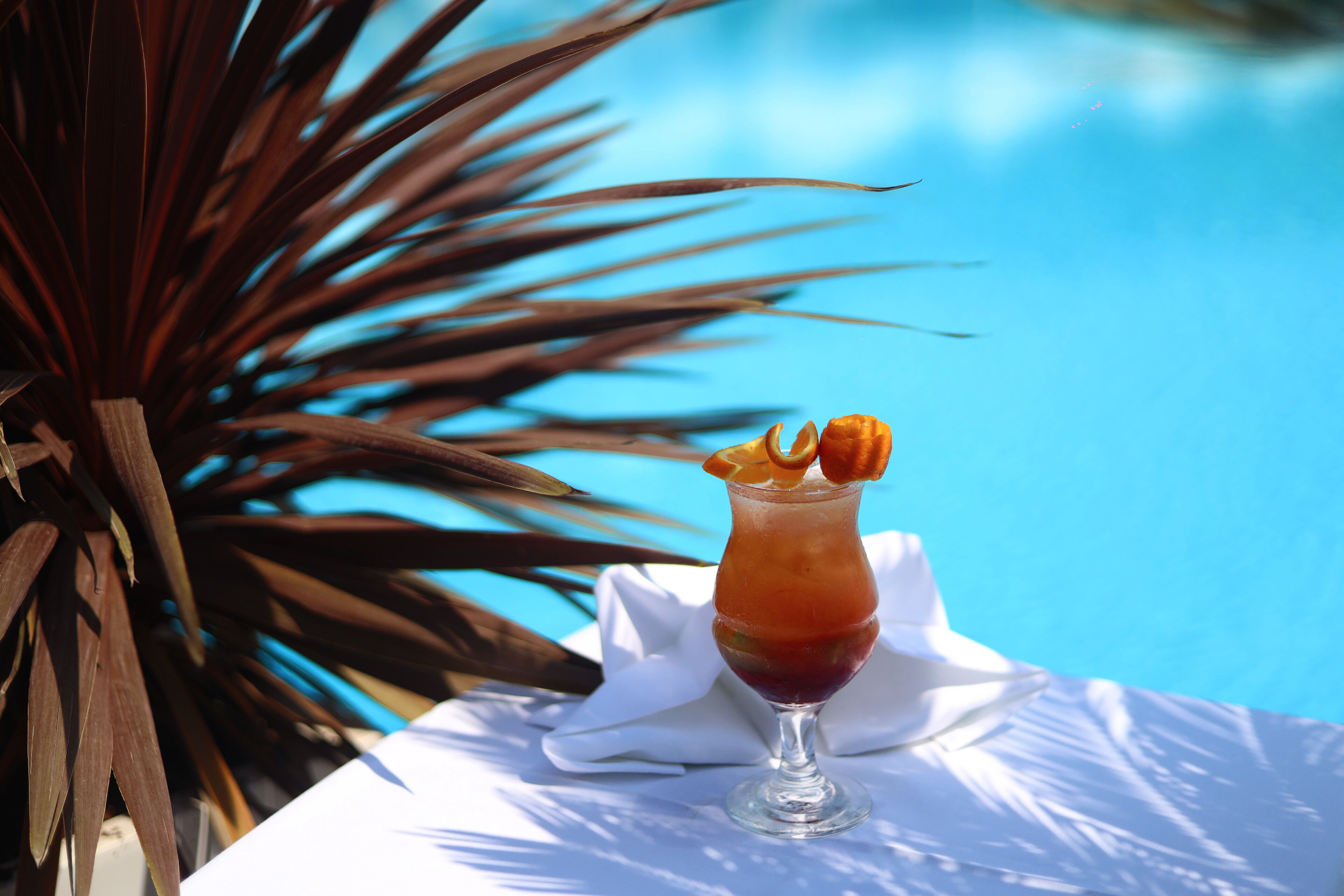 A cocktail on a white table by a pool with a palm plant in the background.