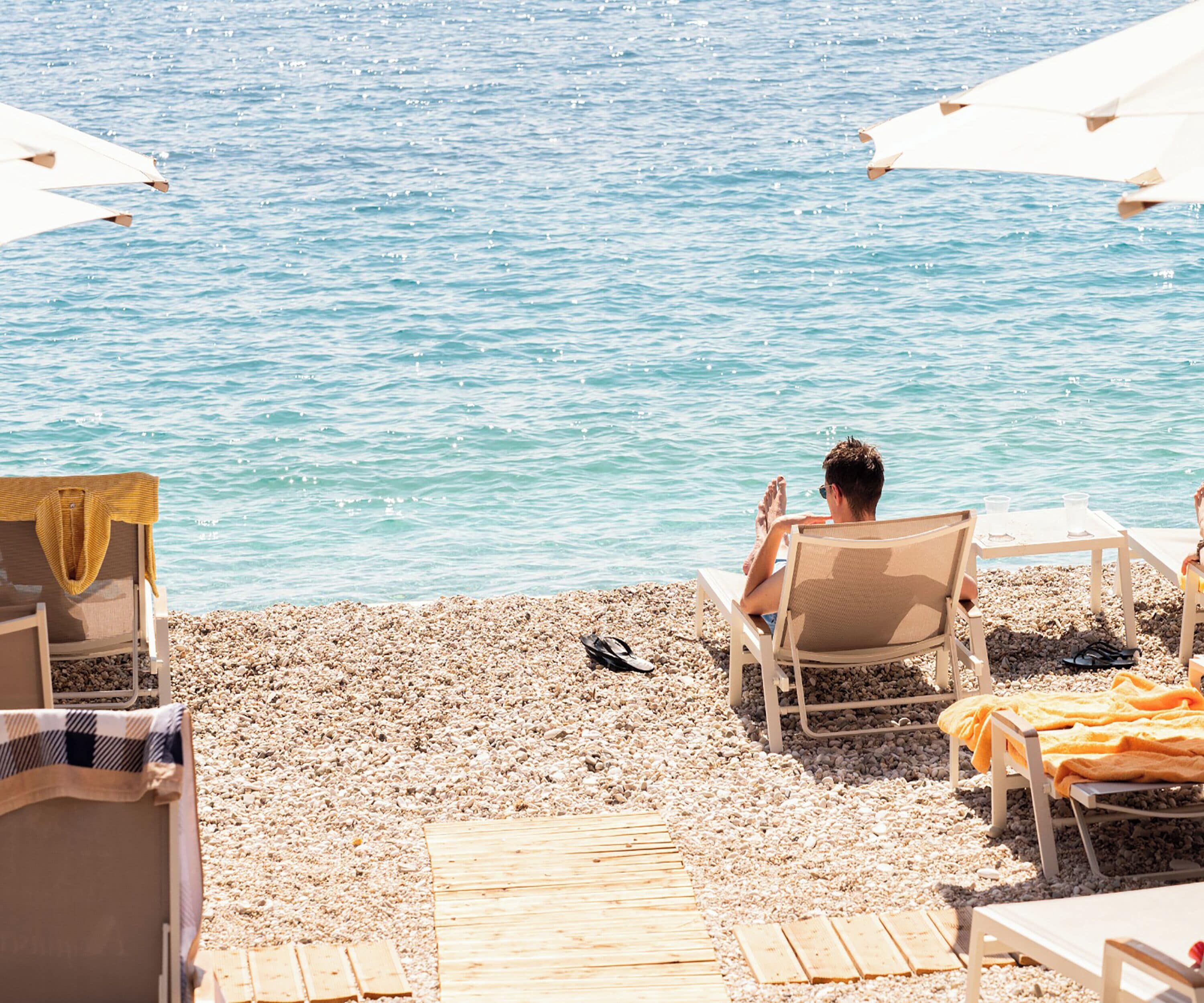 A beach with a man relaxing on a chair, a table with drinks, and umbrellas.