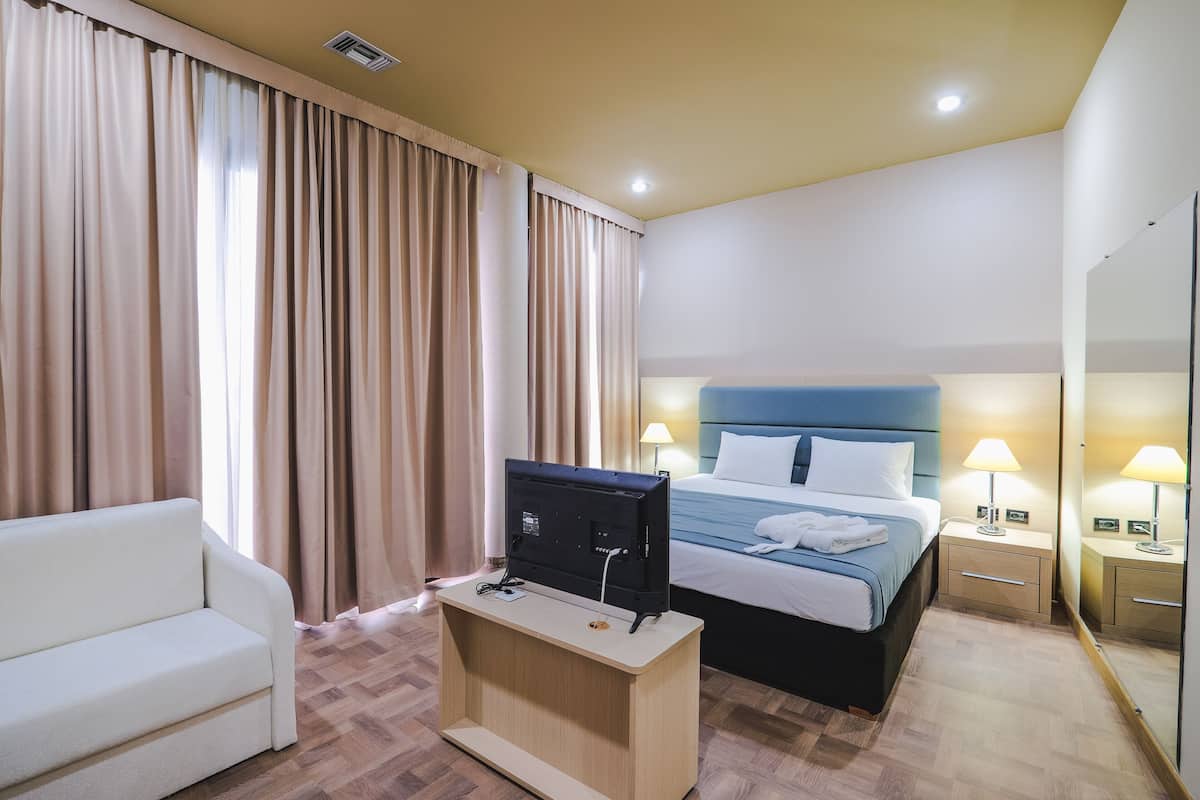 A hotel room with a bed, bedside tables, a TV, a sofa, and curtains.