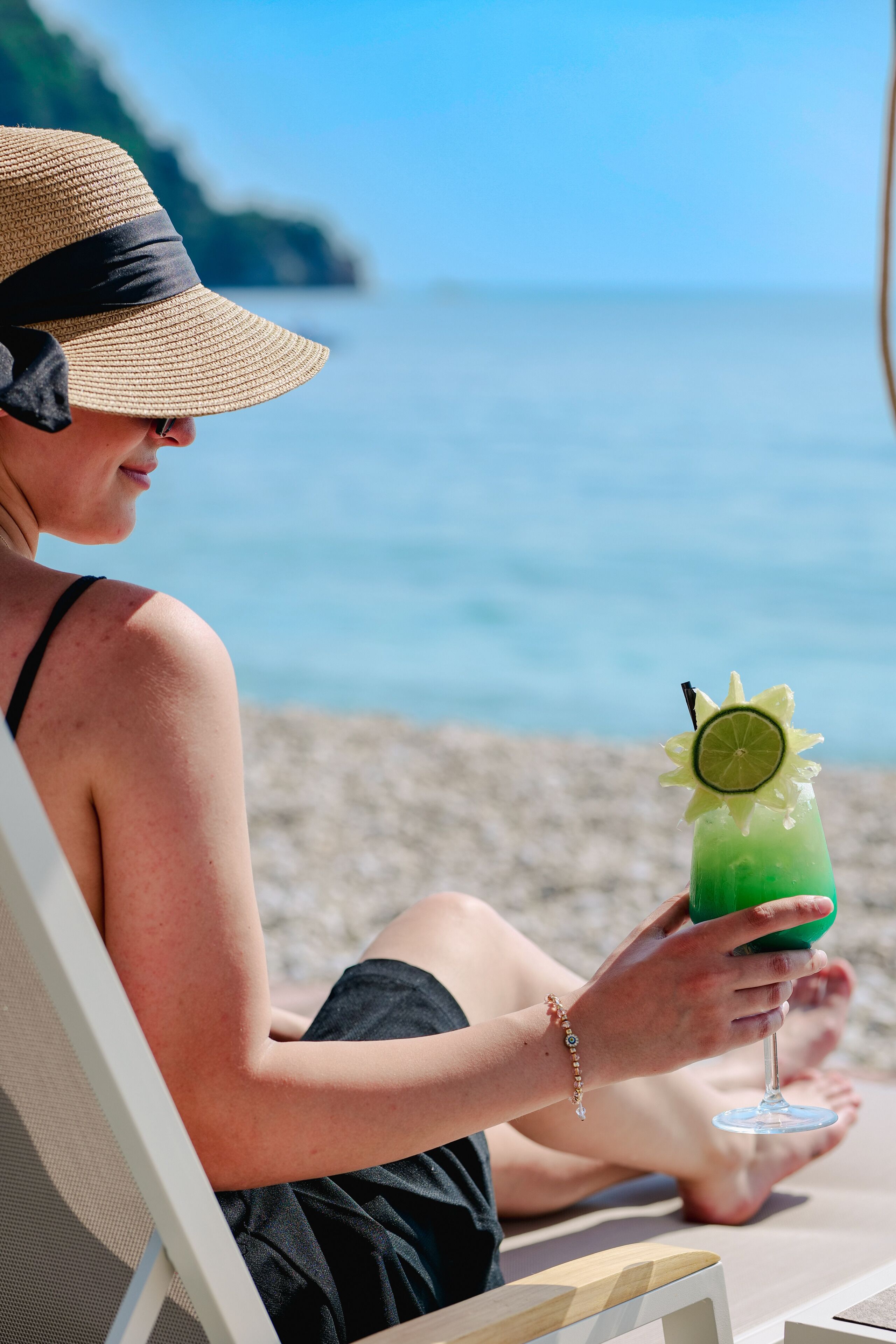 A person sitting on a beach chair holding a green drink with a lime garnish, wearing a straw hat and black swimsuit.