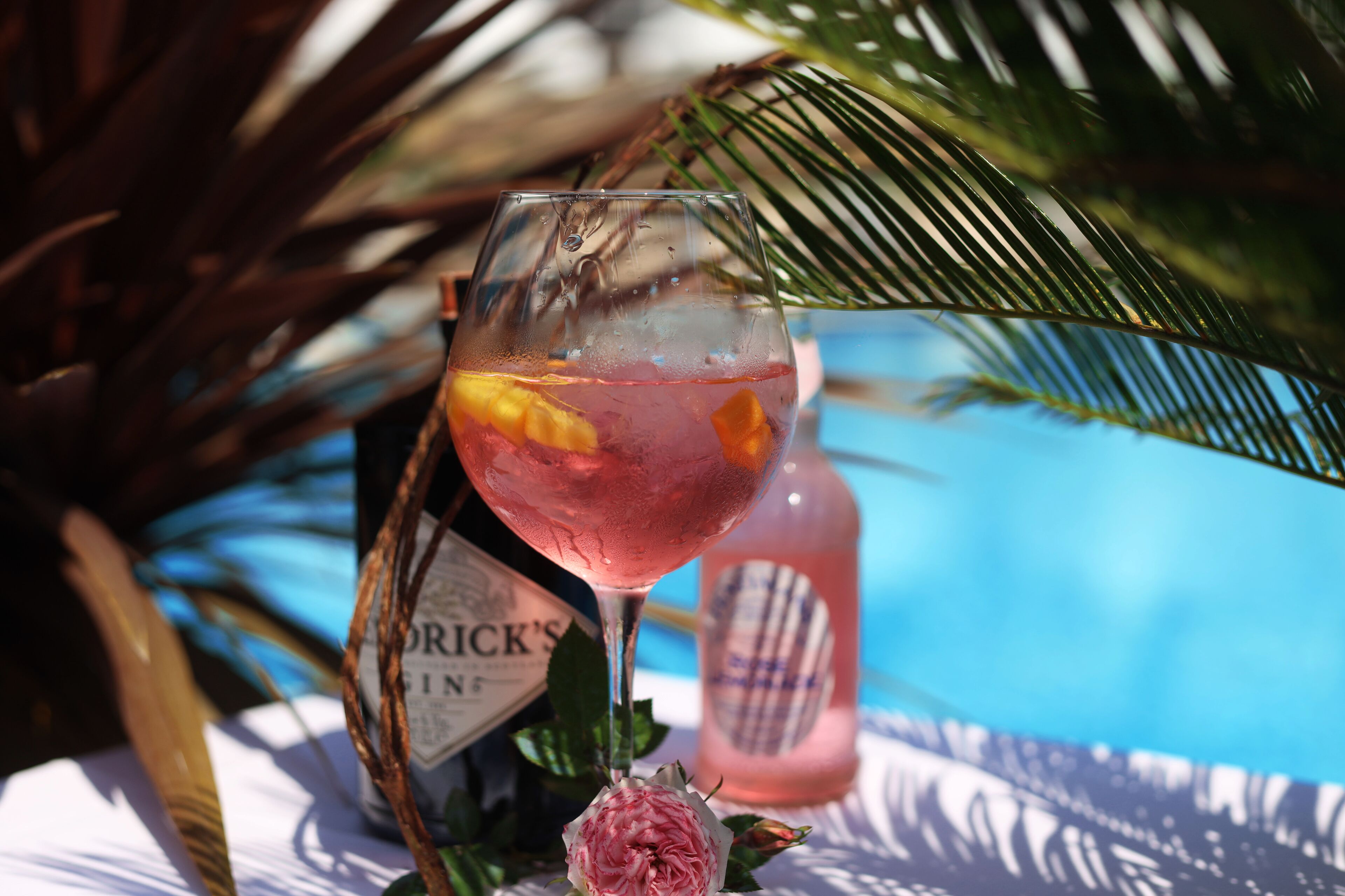 Un got de còctel rosat amb fruita, una ampolla de licor i una piscina al fons.