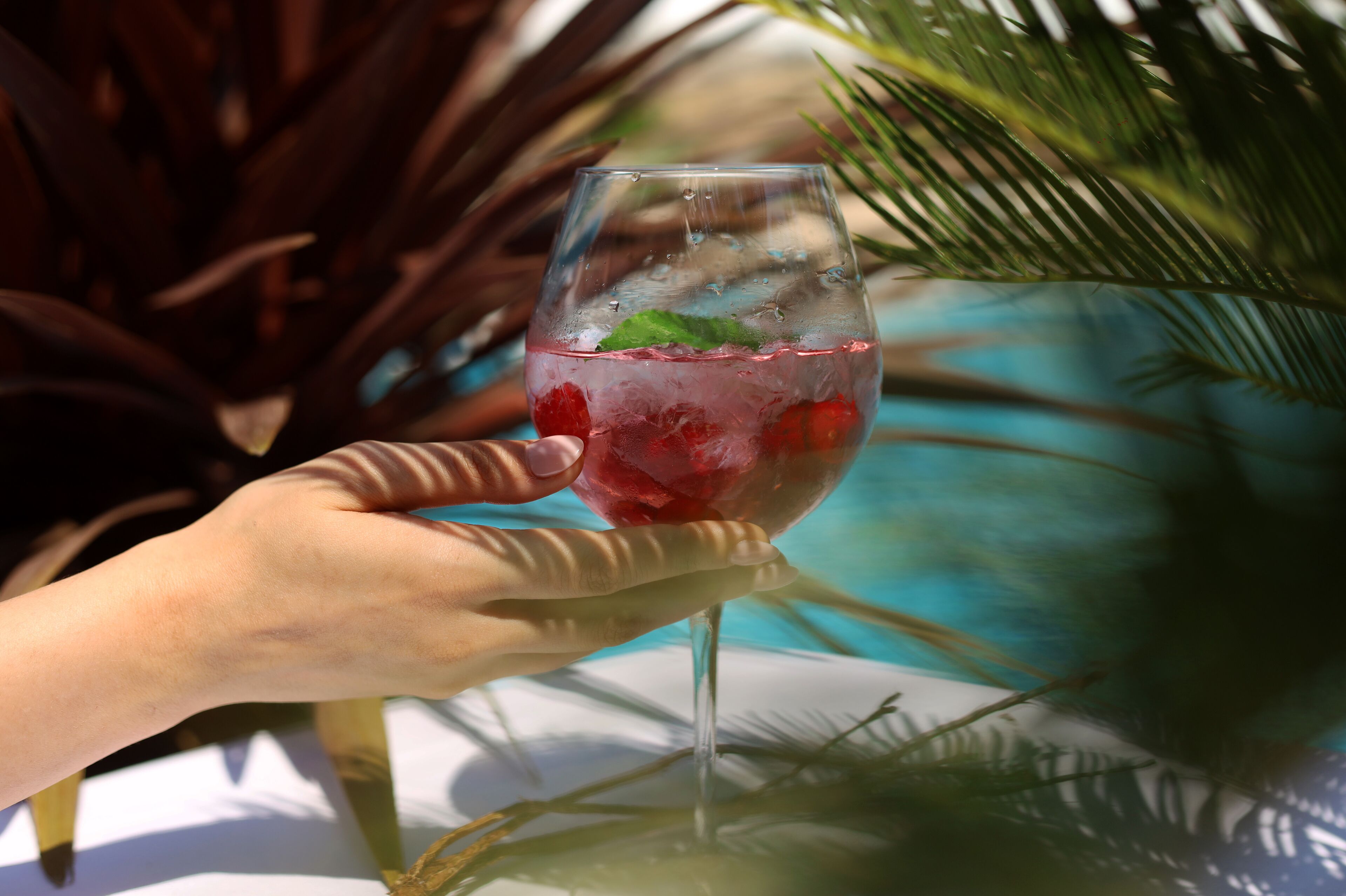 A hand holding a glass with a beverage and ice, near a pool with palm trees.
