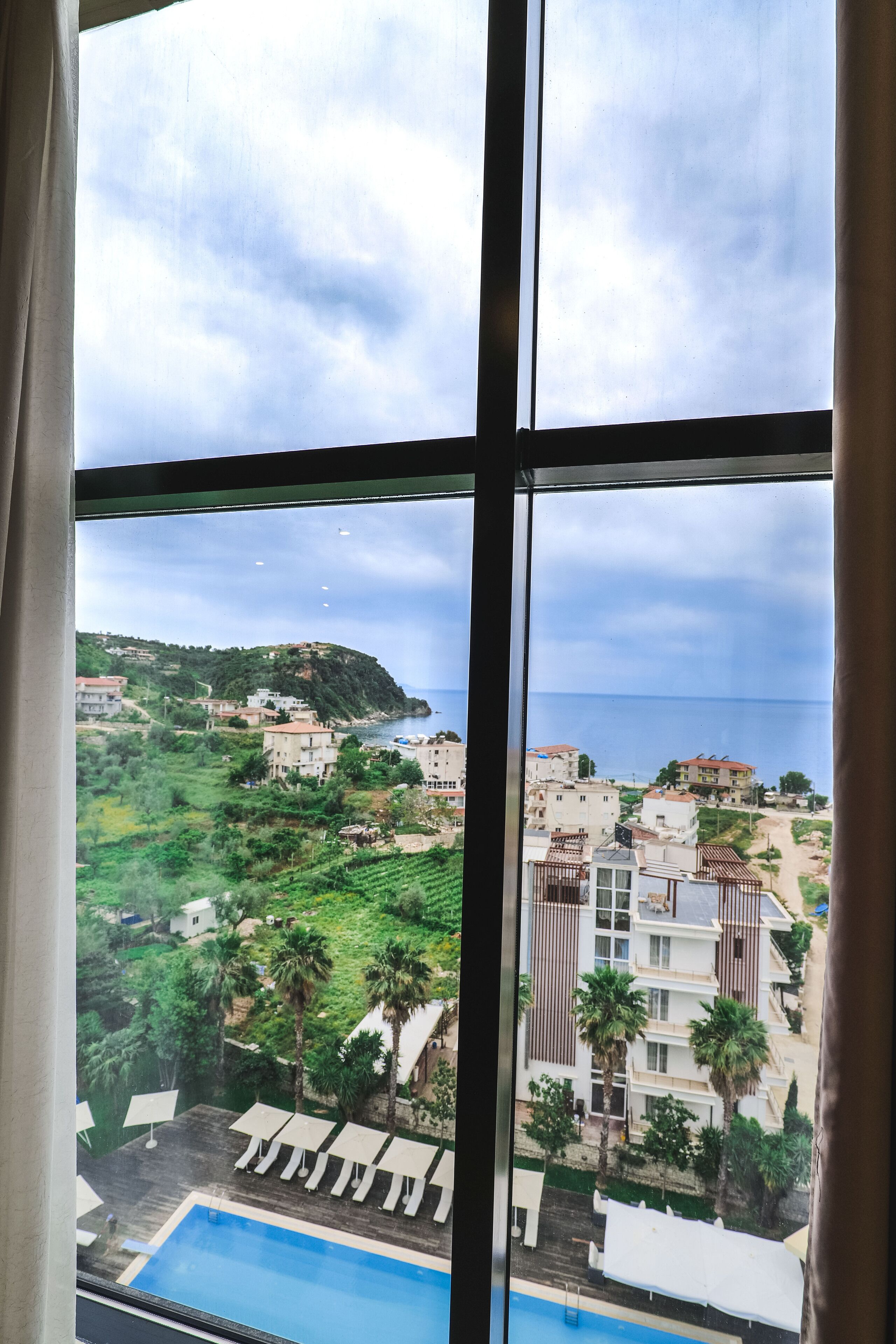 A view from a window overlooking a pool area with lounge chairs, a building with balconies, and a coastal landscape.