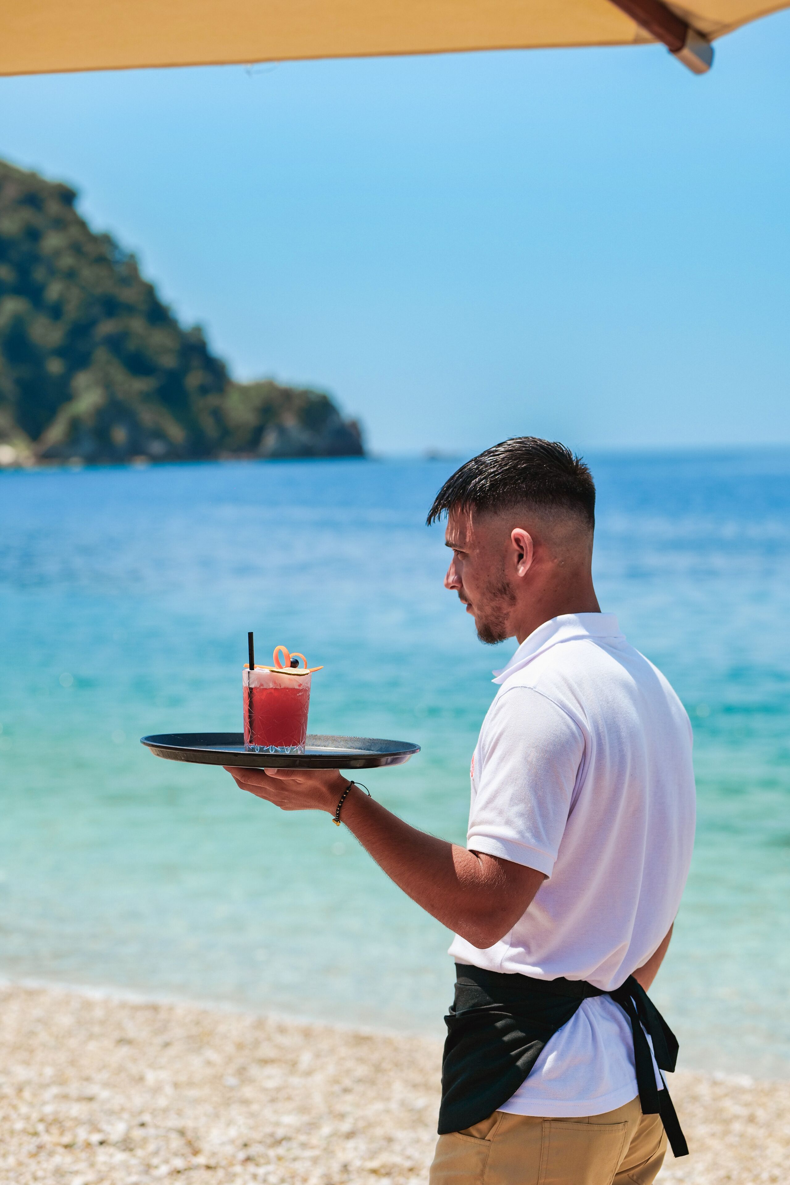 Una persona che tiene un vassoio con una bevanda e una cannuccia, in piedi su una spiaggia con acqua cristallina e una terraferma all'orizzonte.