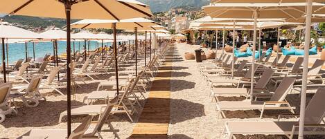 Un resort frente a la playa con hileras de sillas de sol bajo grandes sombrillas, un camino de madera y una vista al mar y montañas al fondo.