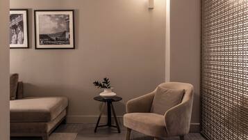 A corner room with a herringbone wooden floor, a beige armchair, a small round table with a plant, and two framed pictures on the wall.