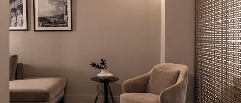 A corner room with a herringbone wooden floor, a beige armchair, a small round table with a plant, and two framed pictures on the wall.