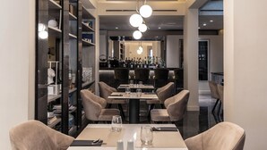 Un espace de restauration moderne avec un bar en arrière-plan, comprenant une table centrale entourée de chaises, une bibliothèque et un luminaire décoratif au plafond.