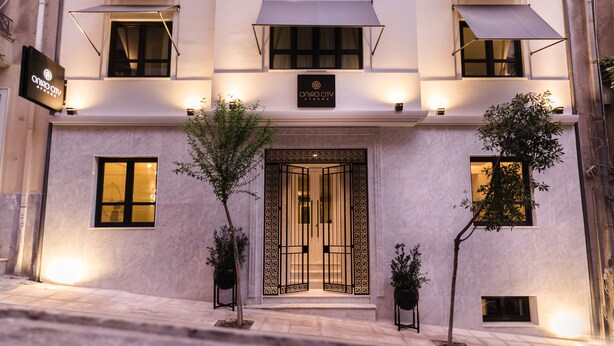 L’entrée d’un hôtel, avec un grand portail orné, une enseigne indiquant « Oniro City Athens », et une façade moderne au design minimaliste.