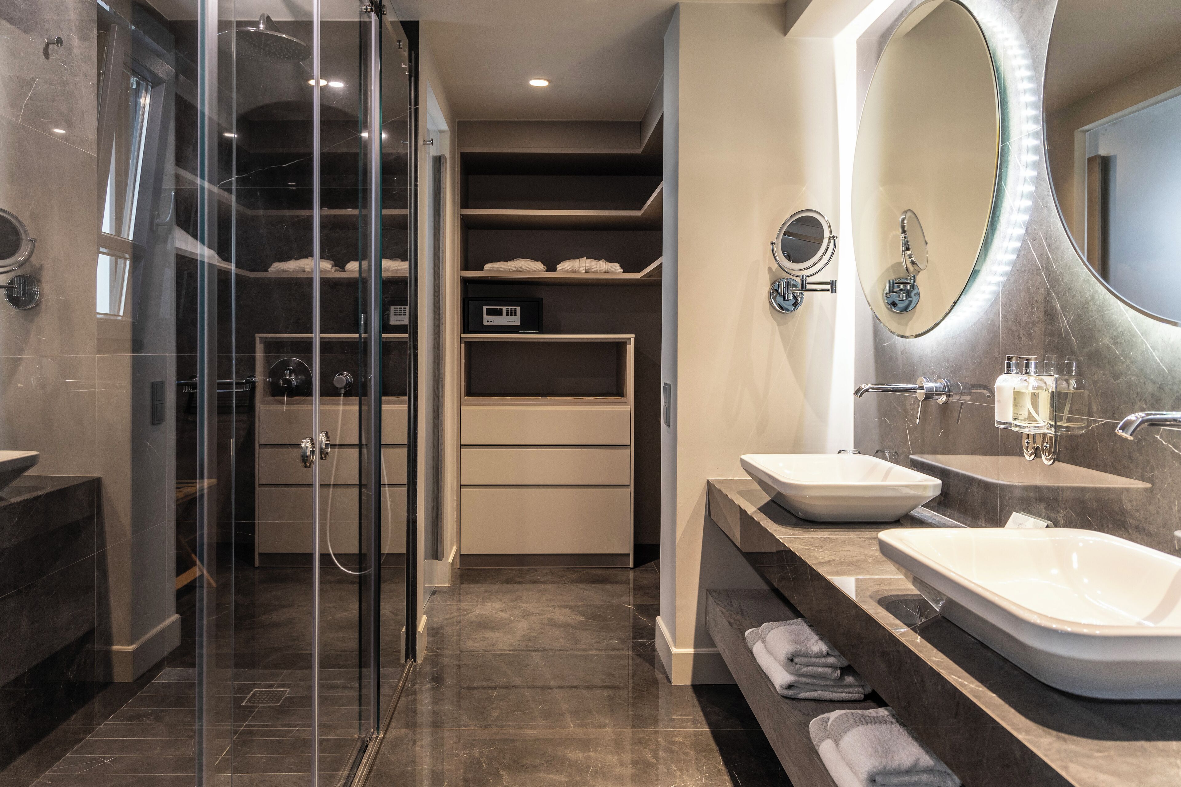 Une salle de bain moderne dotée d’une douche à parois de verre, de deux lavabos, d’un meuble-lavabo avec miroir et d’étagères intégrées.
