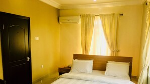 A hotel room with a bed, a nightstand, an air conditioning unit, and a window with curtains. - Milestone Hotel Annex (Lekki)