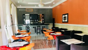 A restaurant with orange and black chairs, a tiled floor, and a kitchen area in the background. - Milestone Hotel Annex (Lekki)