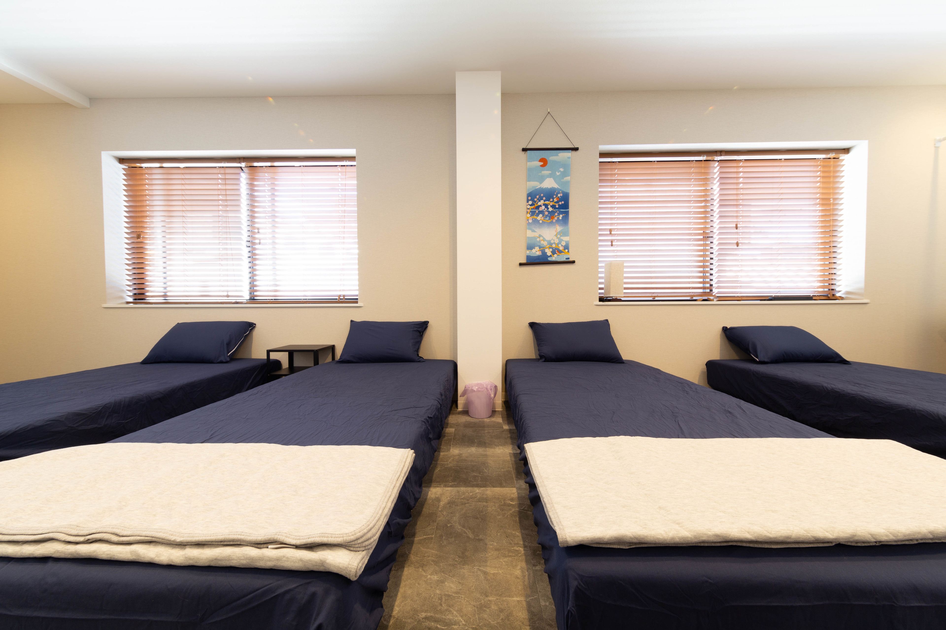 Ein Zimmer mit zwei Betten, jedes mit einem Kopfkissen und einer Decke. Es gibt Fenster mit Jalousien, einen kleinen Tisch und eine Wanddekoration.