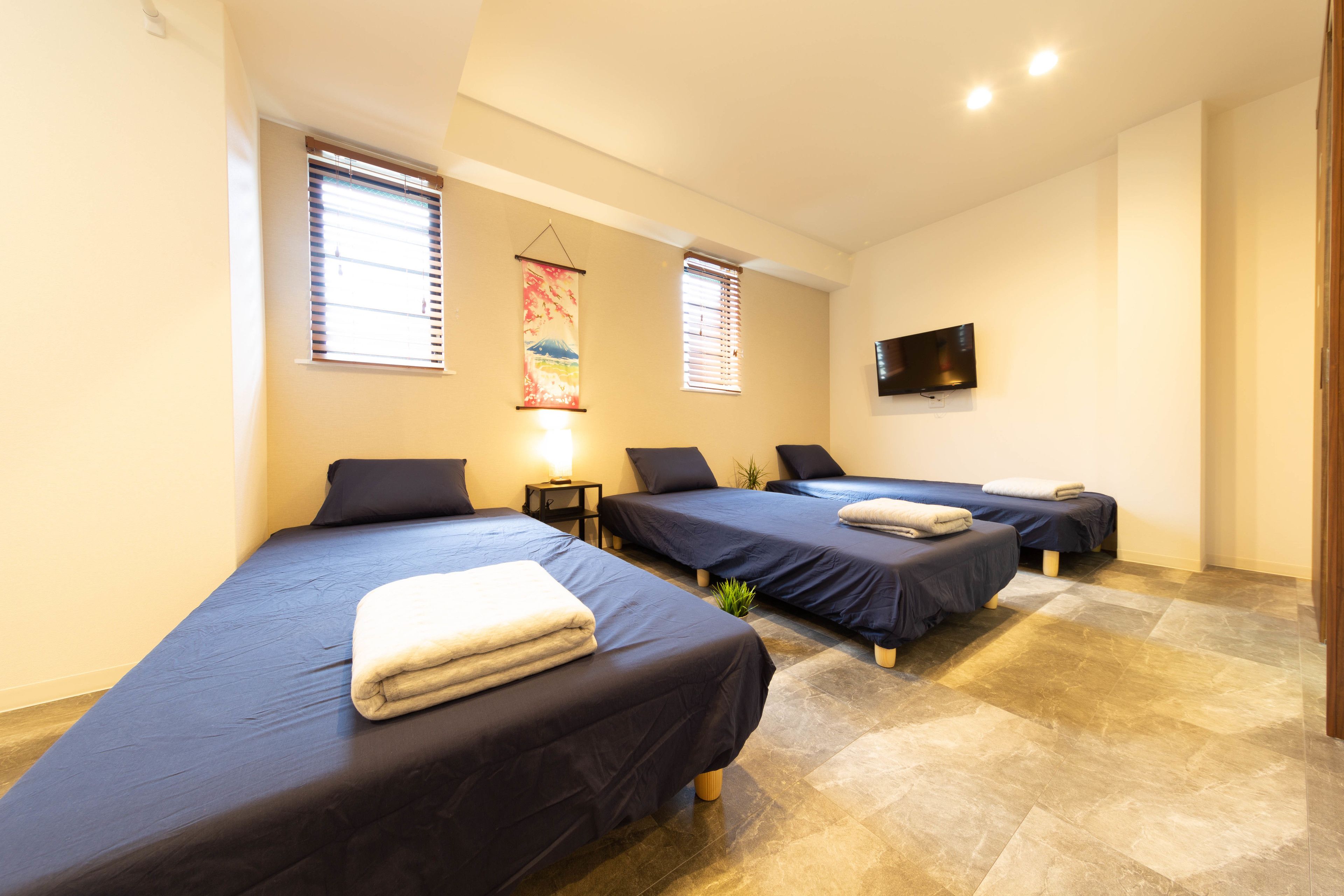 A hotel room with three single beds, a flat-screen TV mounted on the wall, and a window with blinds.
