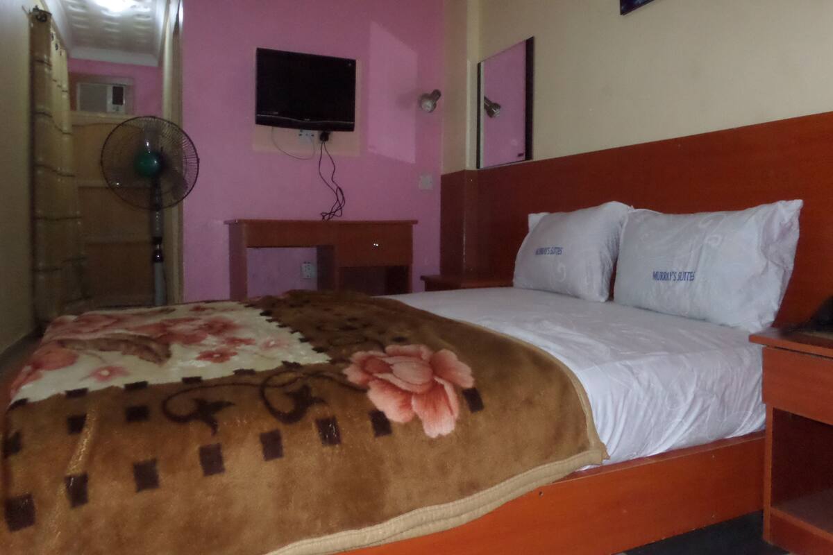 A hotel room with a bed, a TV mounted on the wall, a bedside table, and a fan.