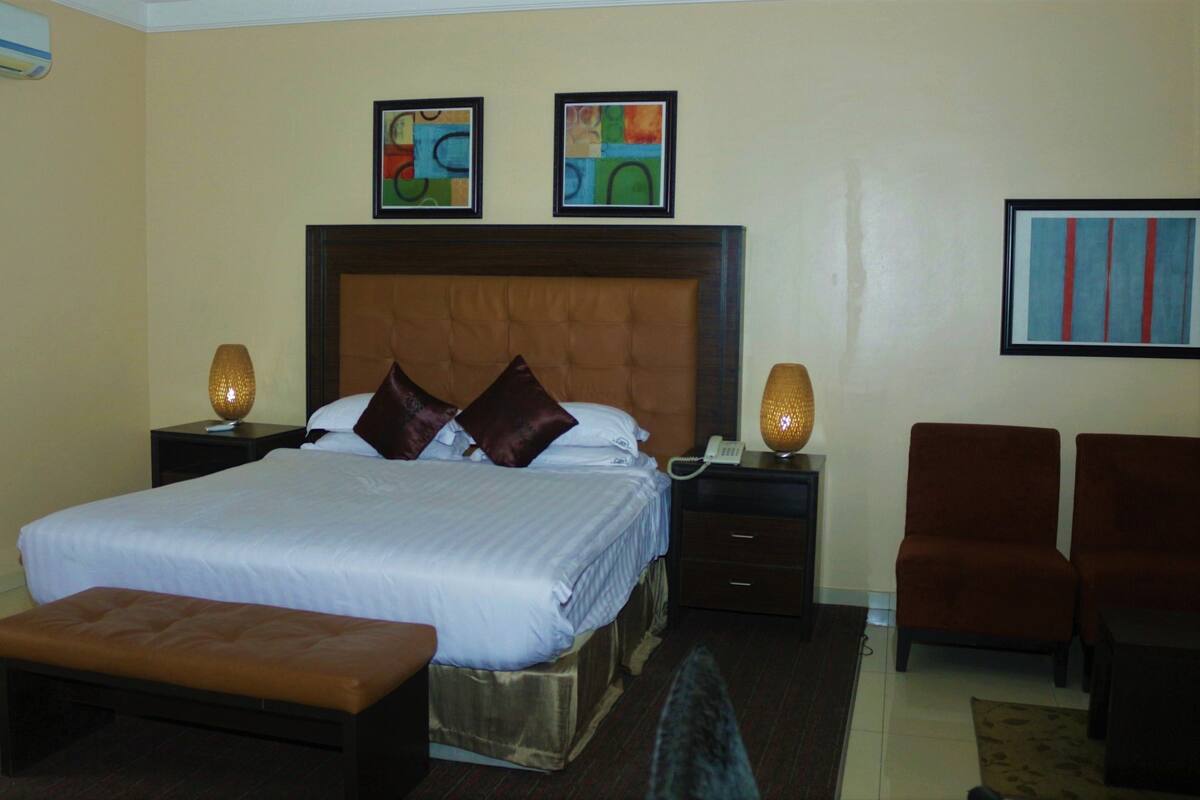 A hotel room with a large bed, two nightstands, a bench, and two chairs. There are two paintings on the wall and an air conditioning unit on the wall.