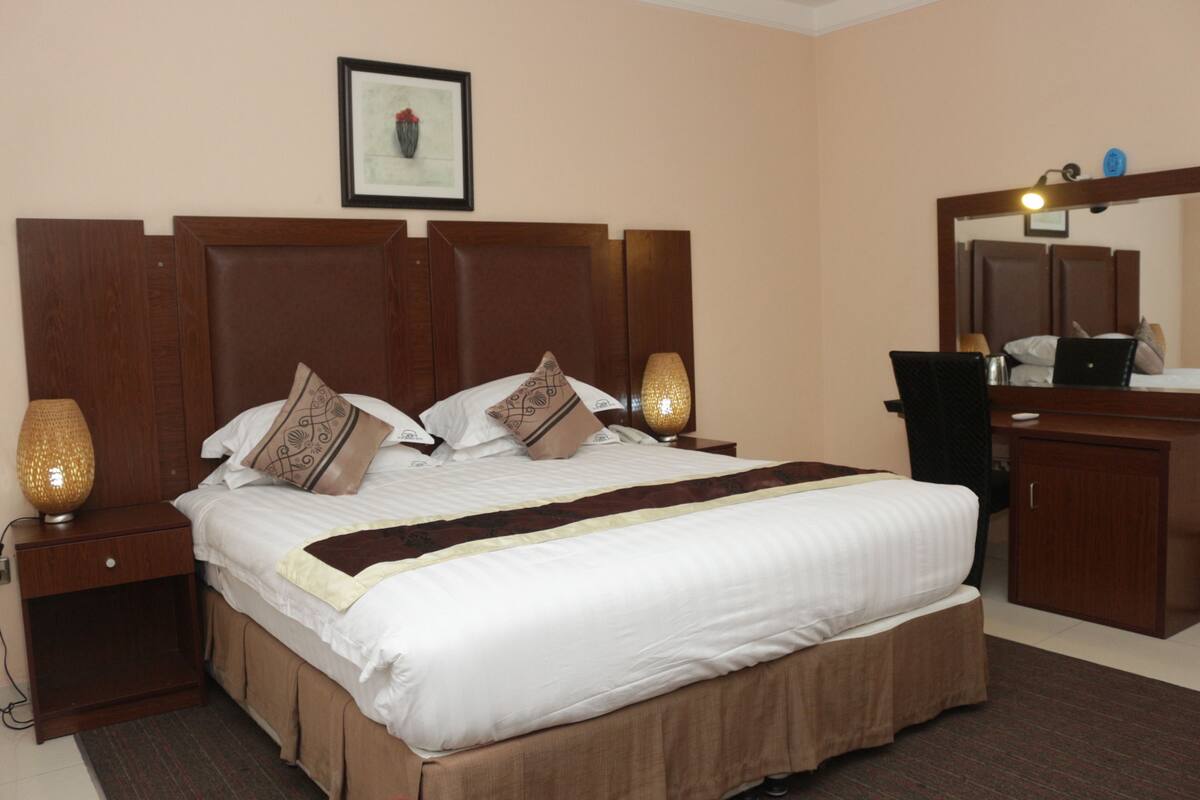 A hotel room with a large bed, a wooden headboard, a bedside table with a lamp, a dresser, and a mirror.
