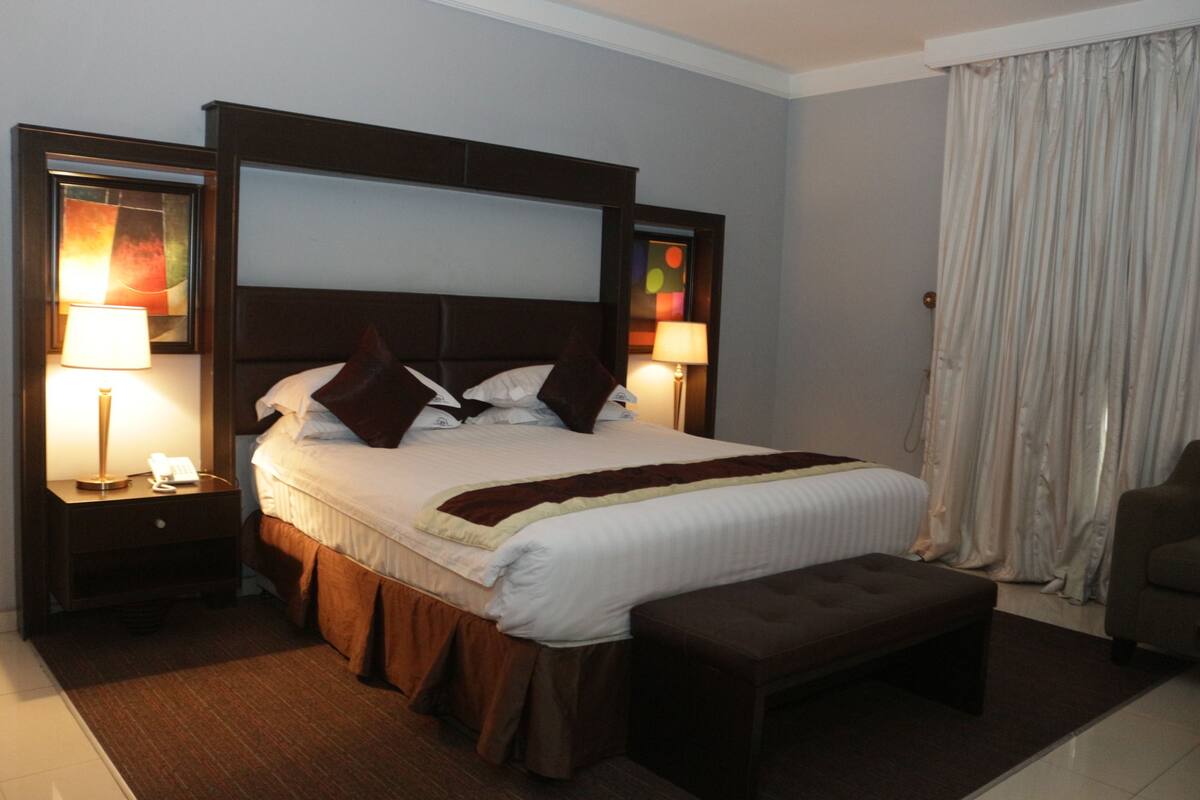 A hotel room with a large bed, two bedside tables, a bench, and a view of the outside through a window with curtains.