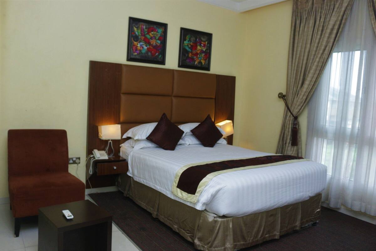 A hotel room with a large bed, a chair, a nightstand, and two paintings on the wall.