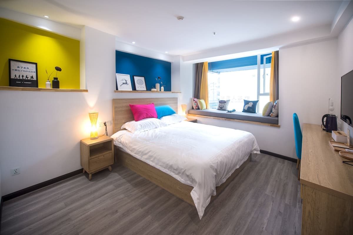 A modern bedroom with a wooden bed, colorful pillows, a bedside table with a lamp, a window with a view, and a desk with a chair.
