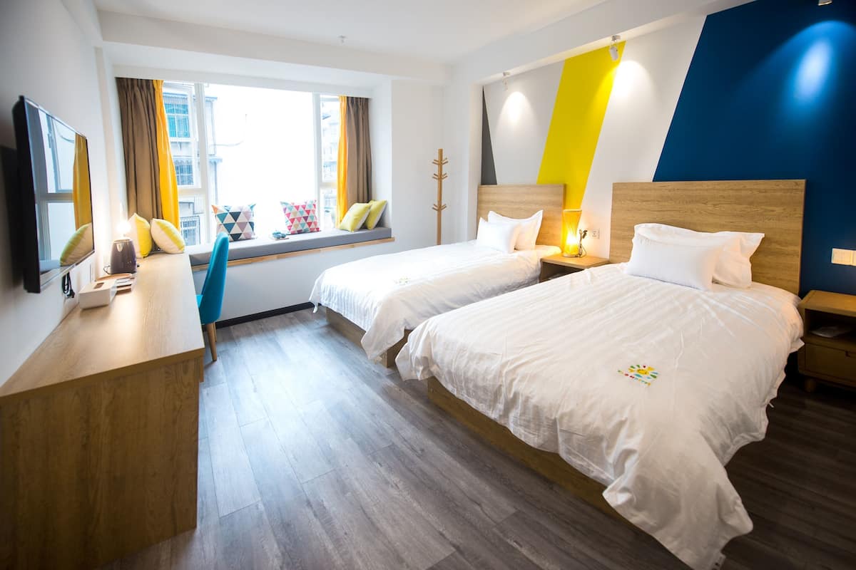 A hotel room with two beds, a wooden desk, a window with curtains, and a view of the city.