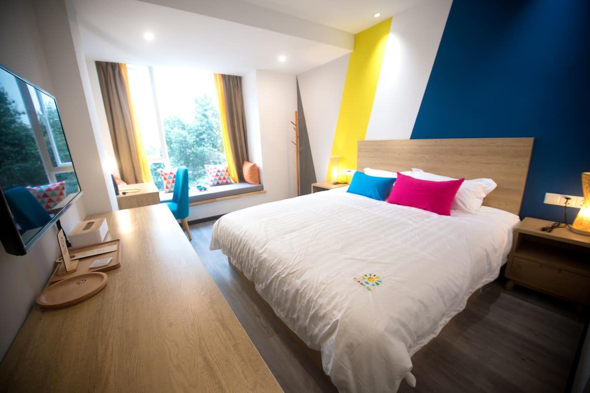 A modern hotel room with a large bed, bedside tables, a flat-screen TV, and a window with curtains.