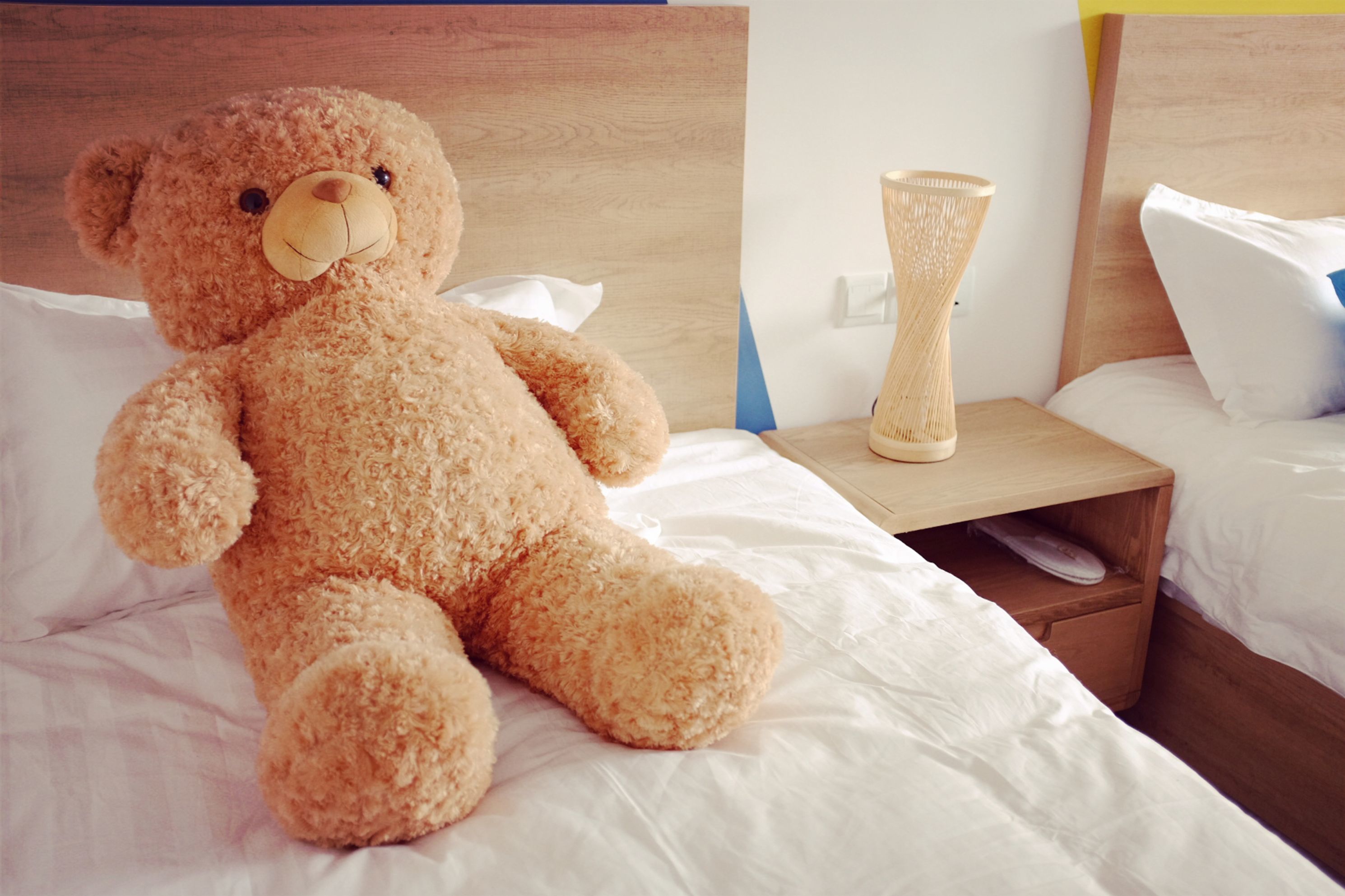 Un oso de peluche grande sobre una cama con sábanas blancas, una mesita de noche de madera con un florero, y una mesita de noche con una lámpara.