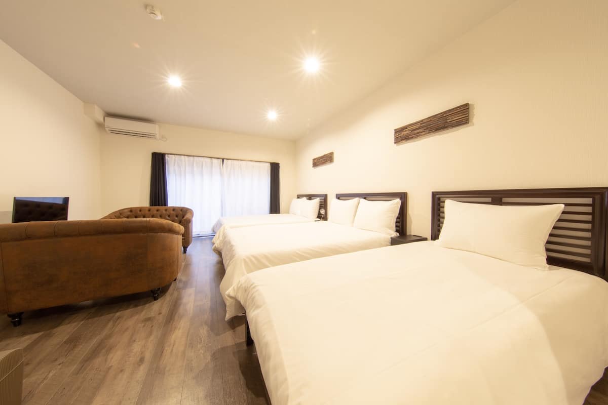 A hotel room with two beds, a sofa, a flat-screen TV, and an air conditioning unit.