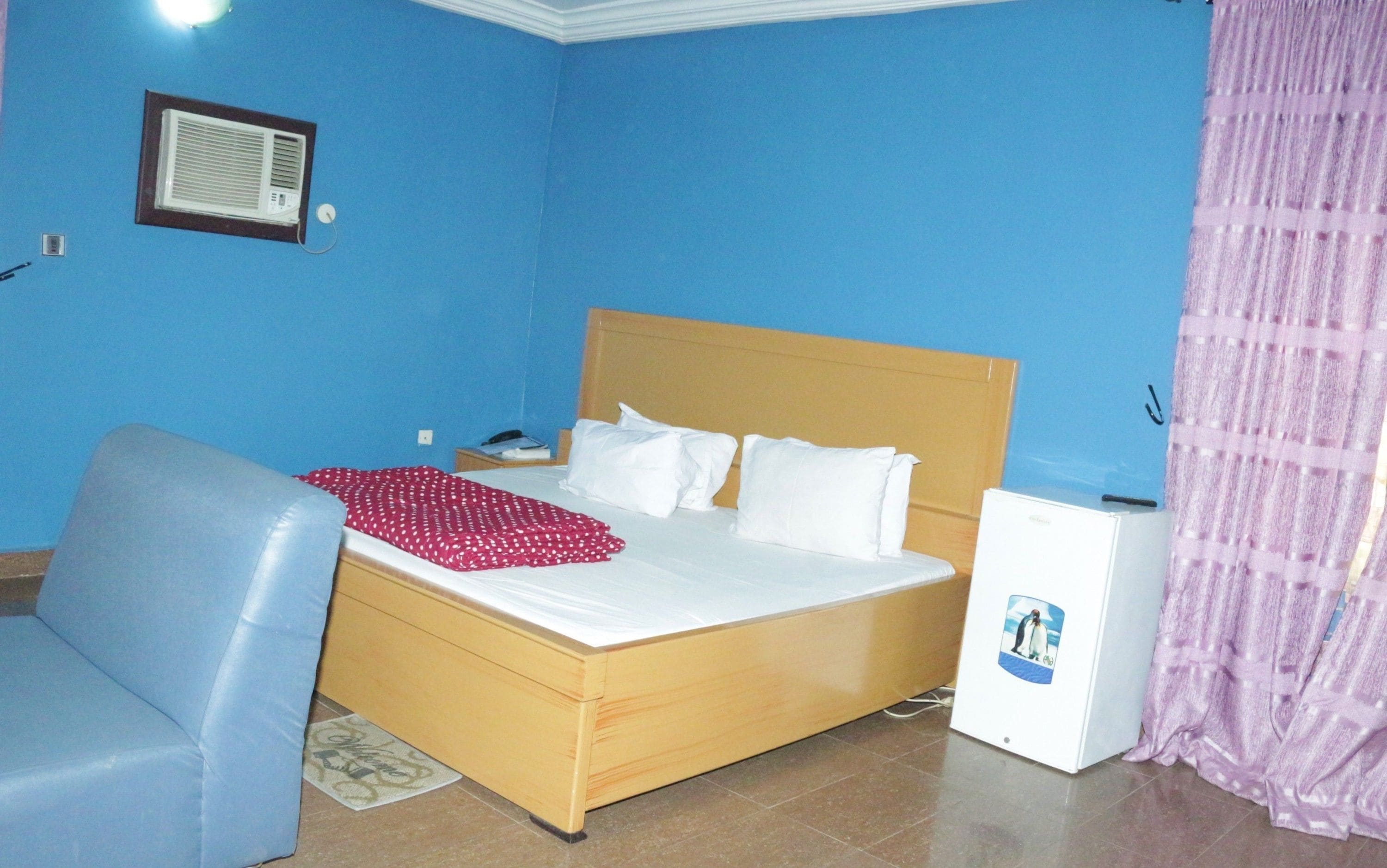 A bedroom with a bed, a desk, a chair, a small refrigerator, and a window with curtains.