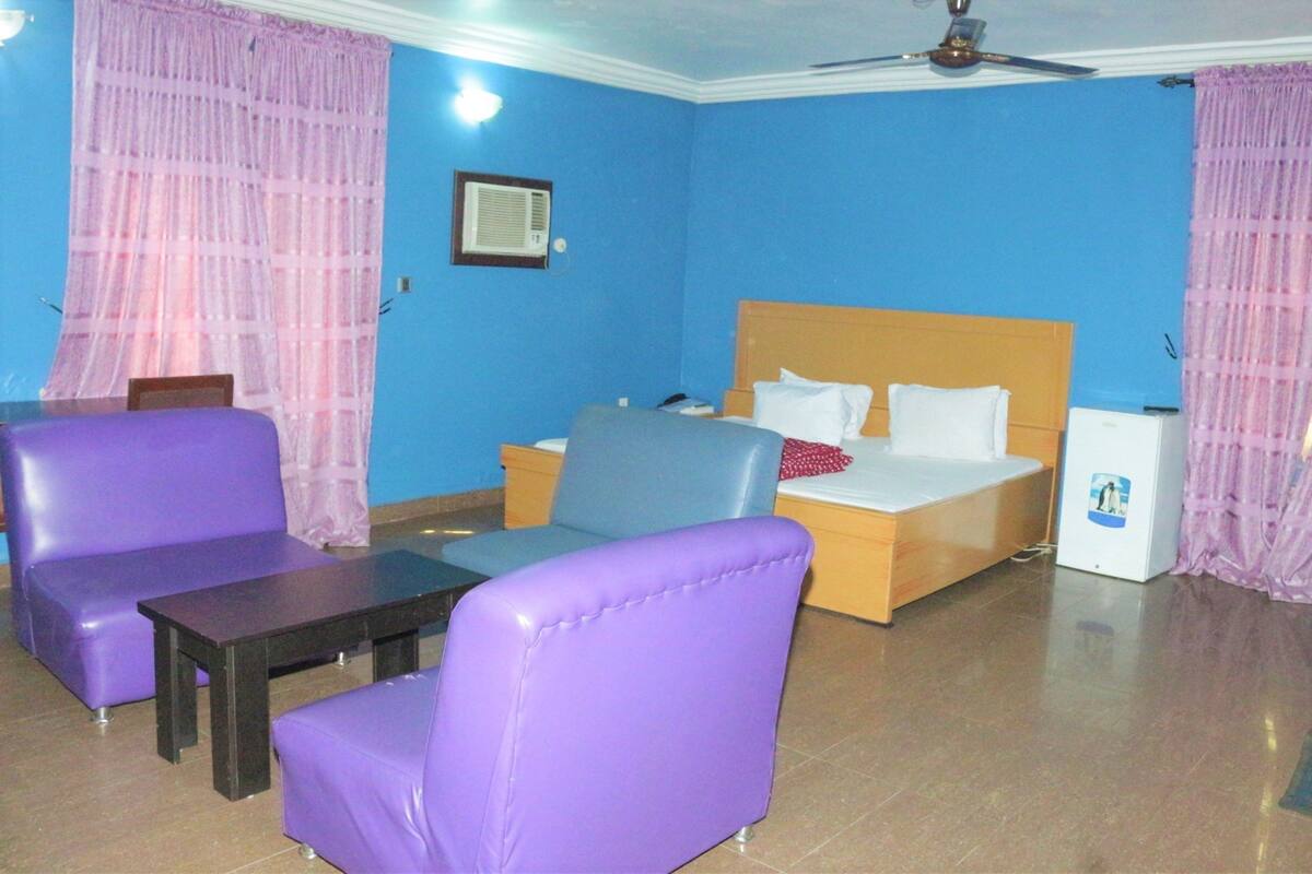 A room with a bed, two purple chairs, a small table, and a ceiling fan.