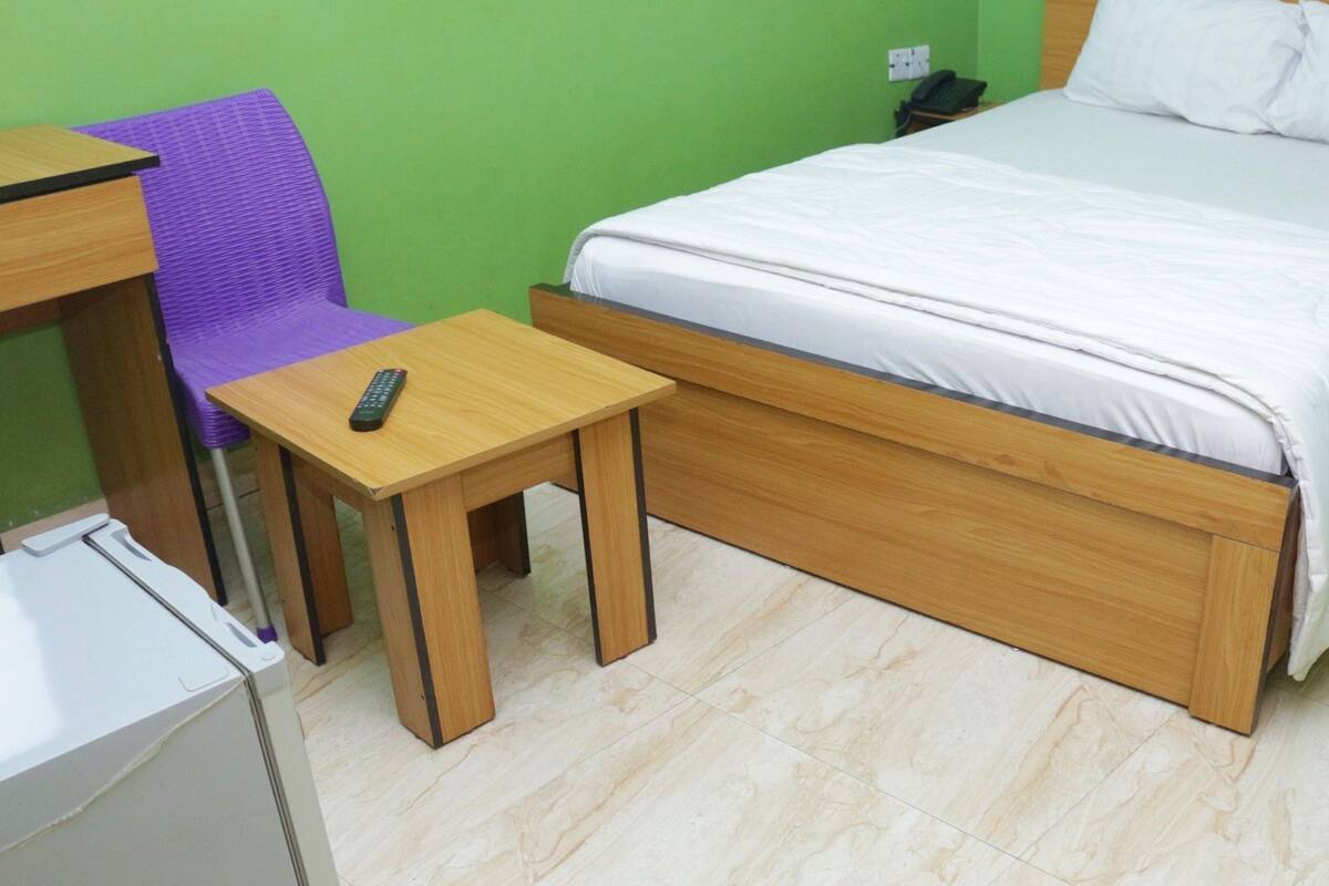 A room with a bed, a small table with a remote control, a purple chair, and a nightstand with a phone.