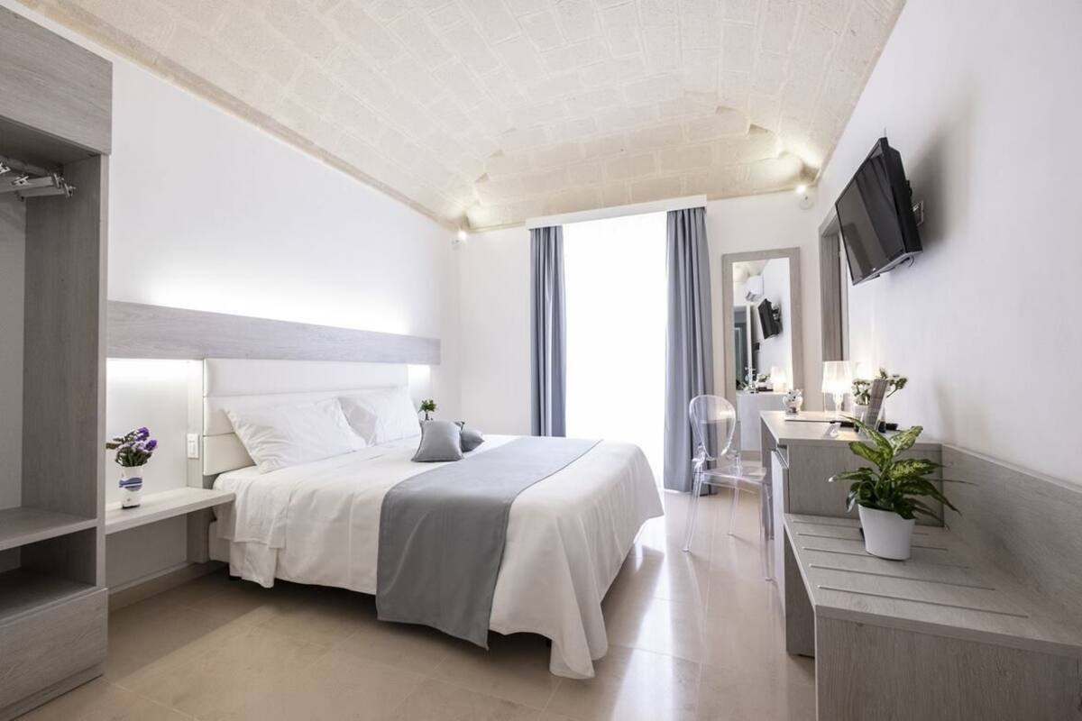 A modern hotel room with a large bed, bedside tables, a desk, a chair, and a TV mounted on the wall.