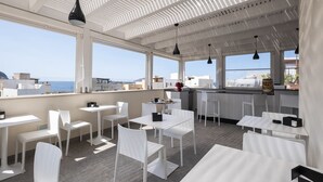 A rooftop dining area with white tables and chairs, large windows offering a view of the sea and buildings, and hanging pendant lights. - Imera B&B (Favignana)