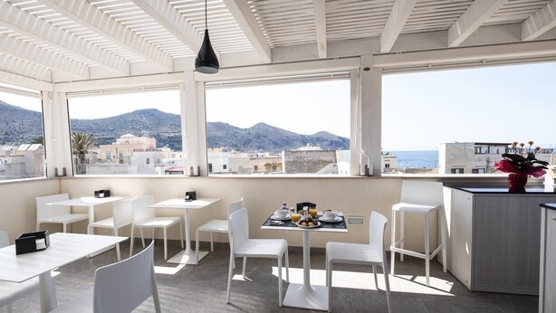 A modern dining area with white furniture and a table set for two, overlooking a scenic mountain view. - Imera B&B (Favignana)