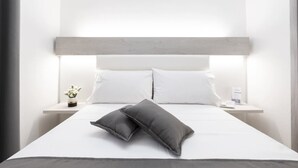A modern hotel room with a minimalist design, featuring a large bed with white bedding and two dark pillows, a bedside table with a small plant, and a nightstand with a lamp. - Imera B&B (Favignana)