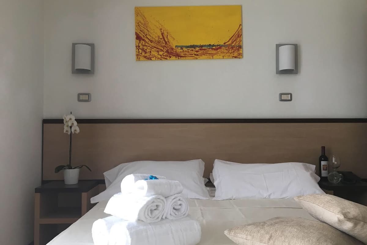 Una habitación de hotel con cama, almohadas, una toalla doblada, mesita de noche con planta, botella de vino y copa de vino.