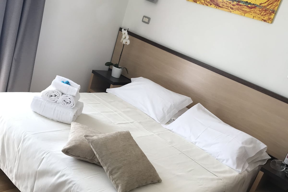 Una cama bien hecha con ropa de cama blanca y un almohadón color beige, una mesita de noche con una orquídea en maceta, y un cuadro en la pared.