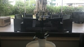 Un espace extérieur abrité, aménagé avec des chaises noires et une table blanche, donnant sur un jardin avec un grand arbre et des bâtiments en arrière-plan.