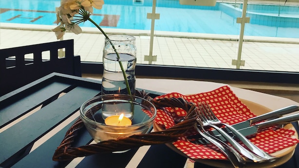 Une table à manger avec une bougie, un verre d’eau et des couverts, placée sur un balcon donnant sur une piscine.