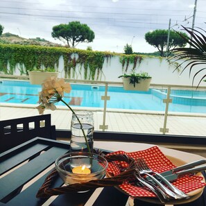 Une table à manger avec une bougie, un verre d’eau et des couverts, placée sur un balcon donnant sur une piscine.