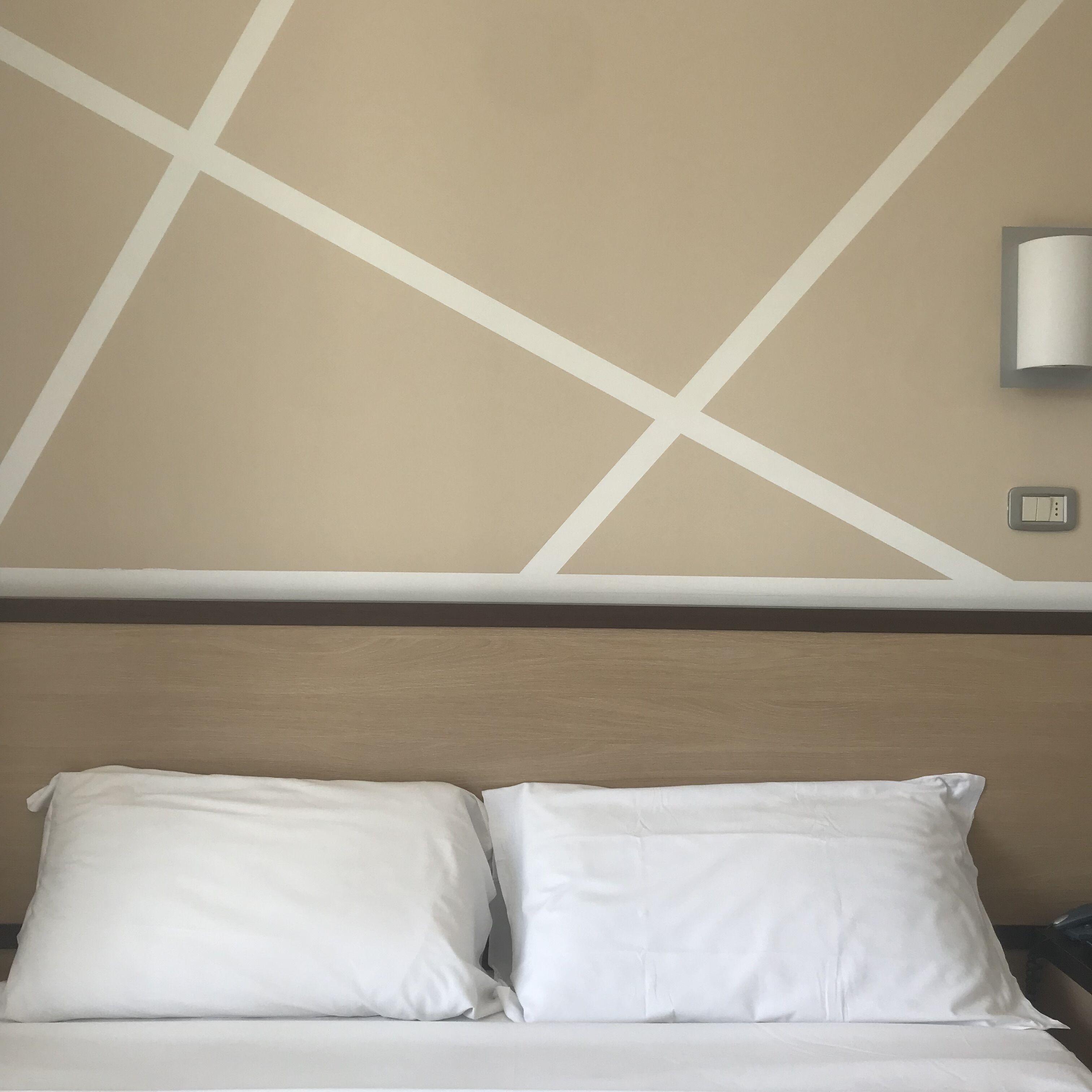 Habitación de hotel con cama, dos almohadas, cabecero y lámpara de pared.
