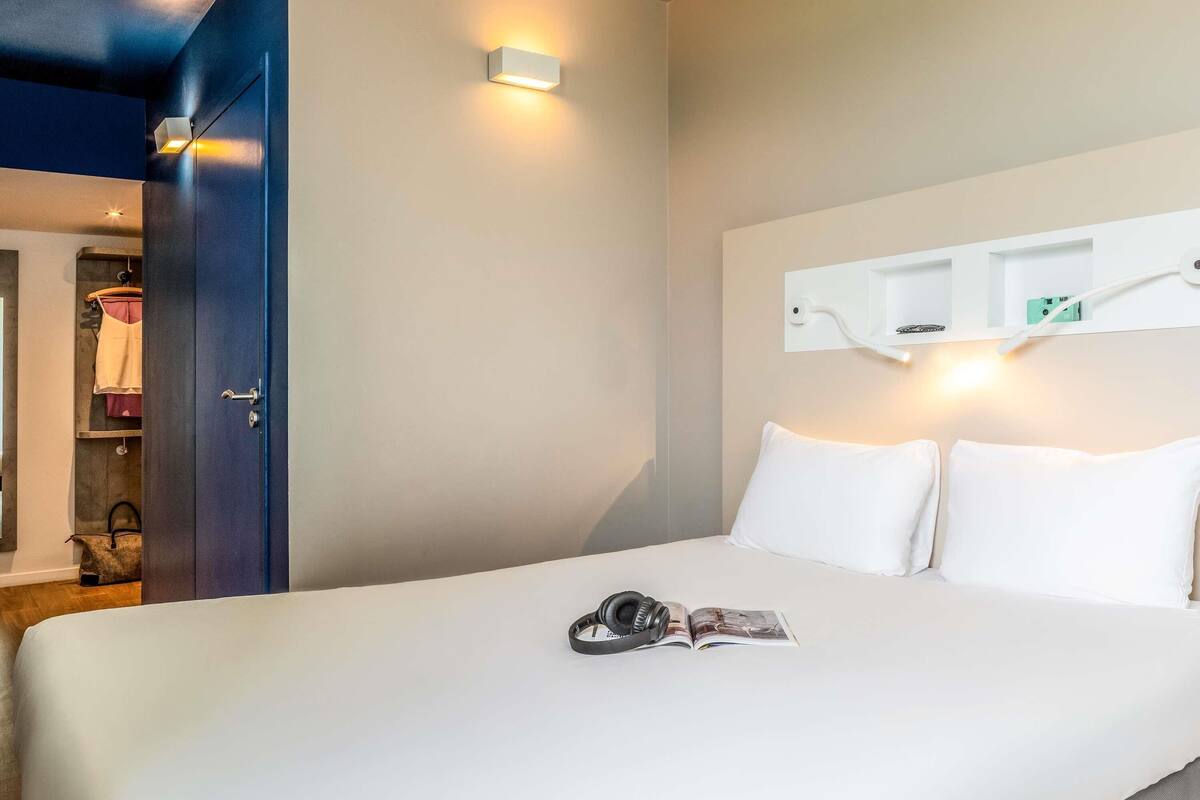 A hotel room with a bed, a nightstand, a wall-mounted shelf, and a door leading to a closet.