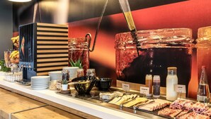 A buffet counter with various food items, a beverage dispenser, and a large poster on the wall. - ibis budget Gonesse Le Bourget (Gonesse)