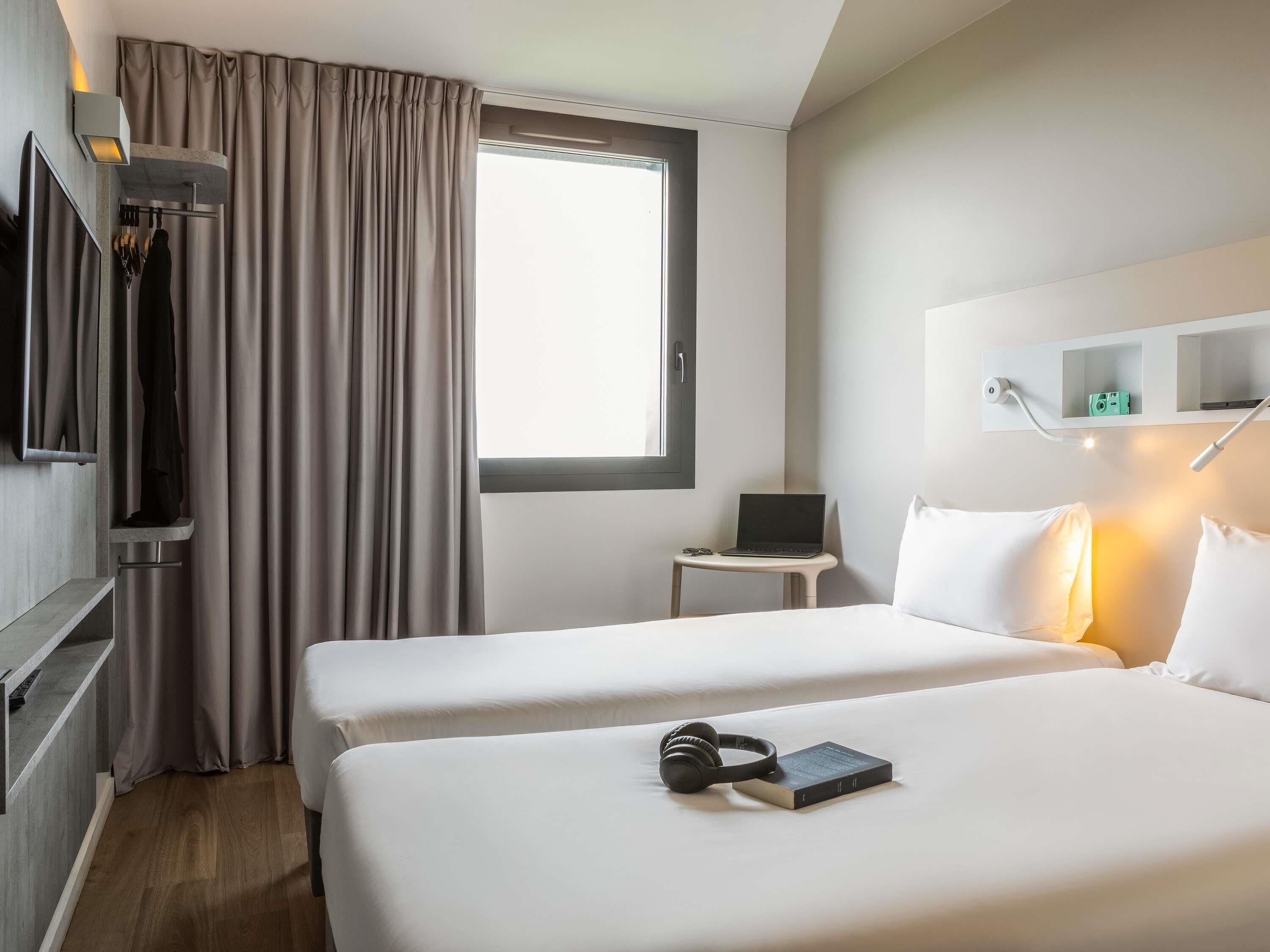 A hotel room with two beds, a desk with a laptop, a TV, and a window with curtains.